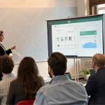 A female founder presents her SaaS project on a projector screen to an engaged audience with laptops, representing a successful launch